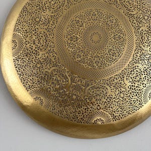 May include: A round, gold-colored metal tray with a decorative, intricate, cut-out design. The design features a repeating pattern of floral and geometric shapes.