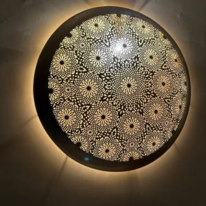 May include: A round, metal ceiling light fixture with a decorative, intricate pattern of cut-out flowers. The light is on and casting a warm glow.