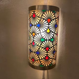 May include: A cylindrical wall sconce with a brass finish and a decorative cut-out pattern. The pattern features a floral design with colorful glass accents in red, blue, yellow, and green. The sconce is mounted on a light-colored wall.