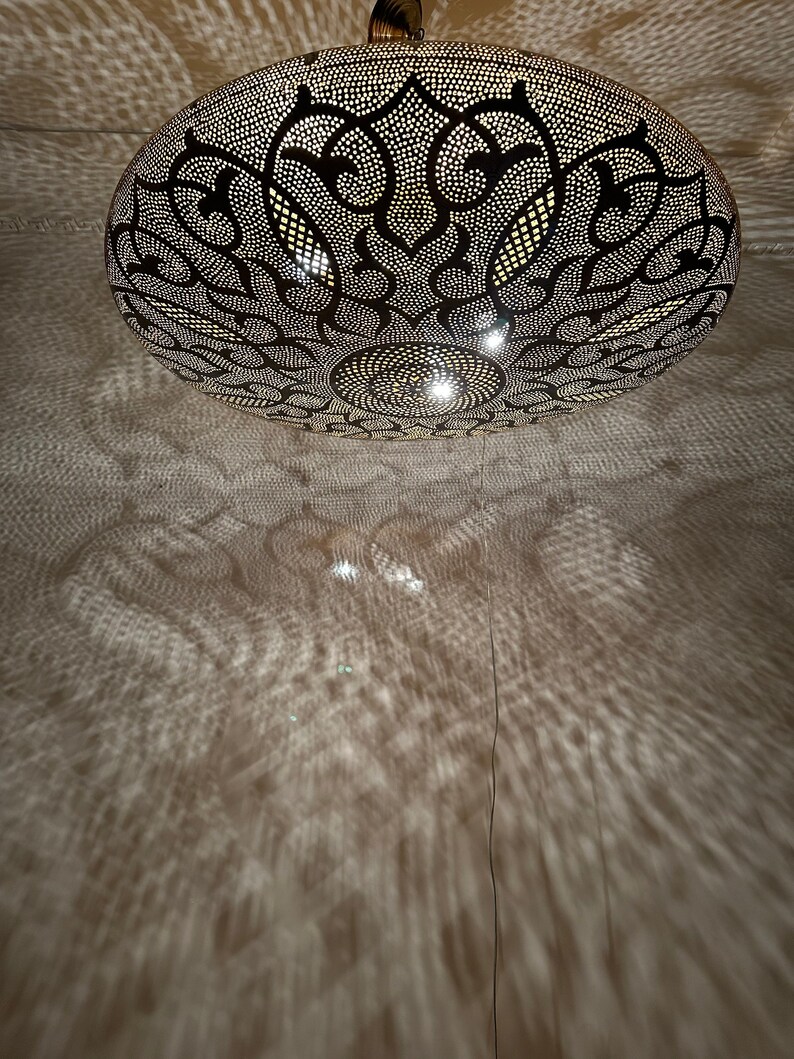 May include: A round, metal ceiling light fixture with a decorative, intricate pattern. The light fixture is made of brass and has a black finish. The light fixture is casting a shadow on the ceiling.