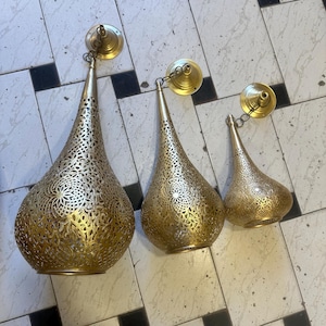 May include: Three gold-colored, teardrop-shaped pendant lights with intricate, openwork patterns. Each light hangs from a chain attached to a circular ceiling mount. The lights vary in size, creating a layered effect.