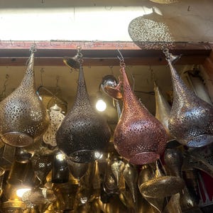 May include: A collection of hanging metal lanterns with intricate, openwork designs. The lanterns vary in color, including gold, bronze, and copper, and are suspended from a wooden beam. The teardrop-shaped lanterns are illuminated from within, creating a warm glow.