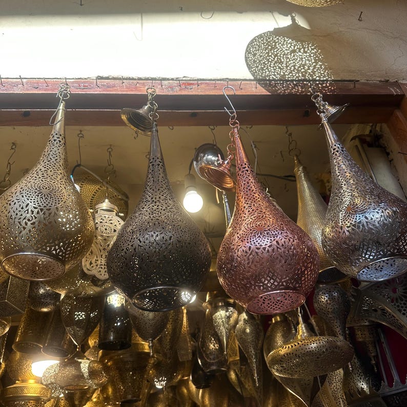 May include: A collection of hanging metal lanterns with intricate, openwork designs. The lanterns vary in color, including gold, bronze, and copper, and are suspended from a wooden beam. The teardrop-shaped lanterns are illuminated from within, creating a warm glow.