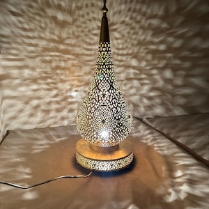 May include: A gold metal table lamp with a decorative, intricate, cut-out design in the shape of a teardrop. The lamp is illuminated and casting a pattern of light on the surface below.