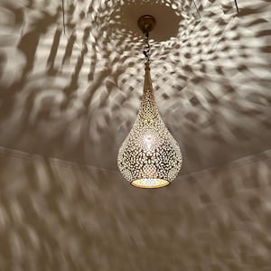 May include: A gold-colored, teardrop-shaped pendant light with intricate cut-out patterns. The light is suspended from the ceiling by a chain and casts decorative shadows. The fixture has a warm, inviting glow, suitable for home decor.