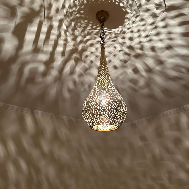 May include: A gold-colored, teardrop-shaped pendant light with intricate cut-out patterns. The light is suspended from the ceiling by a chain and casts decorative shadows. The fixture has a warm, inviting glow, suitable for home decor.