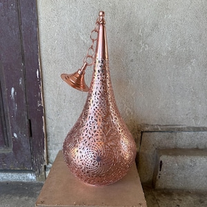 May include: A copper-colored pendant light fixture with an intricate, openwork design. The teardrop-shaped lamp has a pointed top and a decorative chain and canopy. The metal has a warm, reddish-brown hue, and the pattern allows light to filter through.