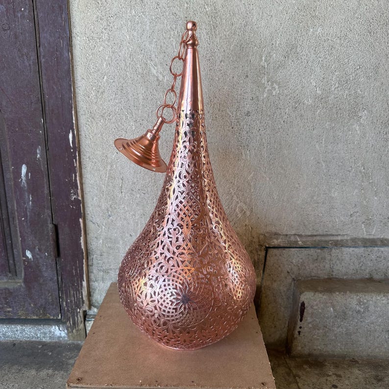 May include: A copper-colored pendant light fixture with an intricate, openwork design. The teardrop-shaped lamp has a pointed top and a decorative chain and canopy. The metal has a warm, reddish-brown hue, and the pattern allows light to filter through.