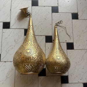 May include: Two gold-colored, teardrop-shaped pendant lights with intricate, openwork patterns. One is attached to a ceiling fixture. The other has a hanging wire. The lights are on a tiled surface.