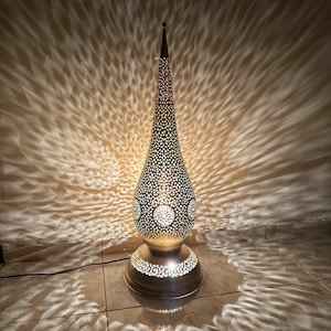 Op de afbeelding: Een gouden staande lamp met een decoratief, ingewikkeld, uitgesneden ontwerp in de vorm van een traan. De lamp is aan en werpt een lichtpatroon op de vloer.