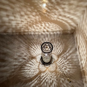 May include: A metal lamp with a decorative, intricate pattern casts a shadow on the floor. The lamp is cylindrical and has a round base.