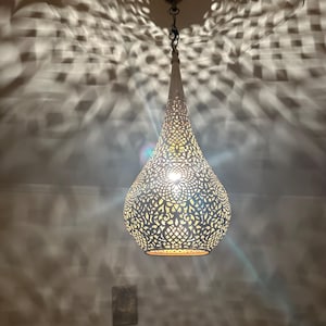 May include: A white, teardrop-shaped pendant light with intricate cut-out patterns. The light fixture is suspended from a chain and casts a patterned shadow on the wall. The interior glows with warm light, creating a decorative ambiance.