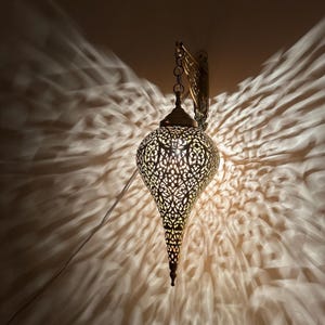 以下が含まれることがあります： 真鍮色の装飾的な壁掛け照明器具。 涙の形をしたシェードには、複雑な切り抜き模様があり、壁に装飾的な光のパターンを投影します。 チェーンがシェードを壁掛けに取り付けています。
