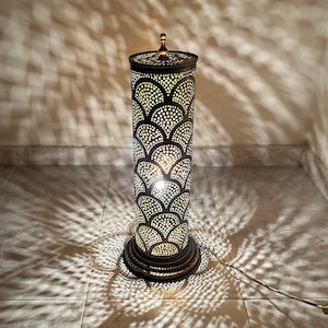 May include: A tall, cylindrical floor lamp with a decorative, intricate metal design. The lamp is made of brass and has a warm, golden finish. The design features a repeating pattern of overlapping circles and arches, creating a delicate and elegant look. The lamp is lit, casting a soft, warm glow.