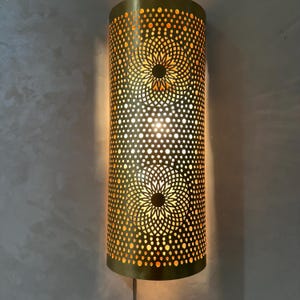 May include: A cylindrical, gold-colored wall sconce with a perforated design. The sconce features two floral patterns and emits a warm, inviting light. The design is a decorative lighting fixture.