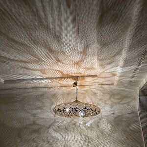 May include: A gold-toned metal pendant light fixture with a round, openwork design casts intricate shadows on the ceiling. The light fixture has a chain and a decorative finial.