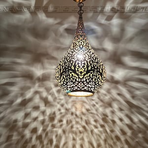 May include: A decorative pendant light with a metal, intricate, cut-out design in the shape of a teardrop. The light is on and casting a pattern of light and shadow on the ceiling.
