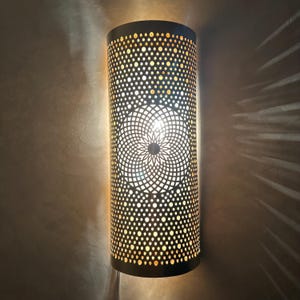 Könnte beinhalten: Zylindrische Wandleuchte aus dunklem Metall. Sie hat ein Muster aus kleinen und großen Perforationen mit einem zentralen floralen Design. Licht scheint durch die Löcher und erzeugt ein warmes Leuchten und dekorative Schatten. Die Leuchte ist ca. 30 cm hoch.