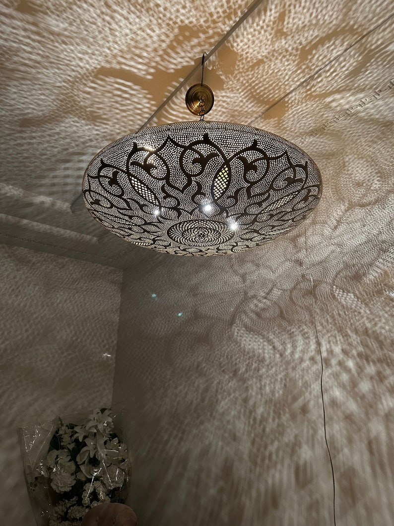 May include: A large, round, metal ceiling light fixture with intricate cut-out designs. The light fixture is hanging from the ceiling and casting a patterned shadow on the wall.