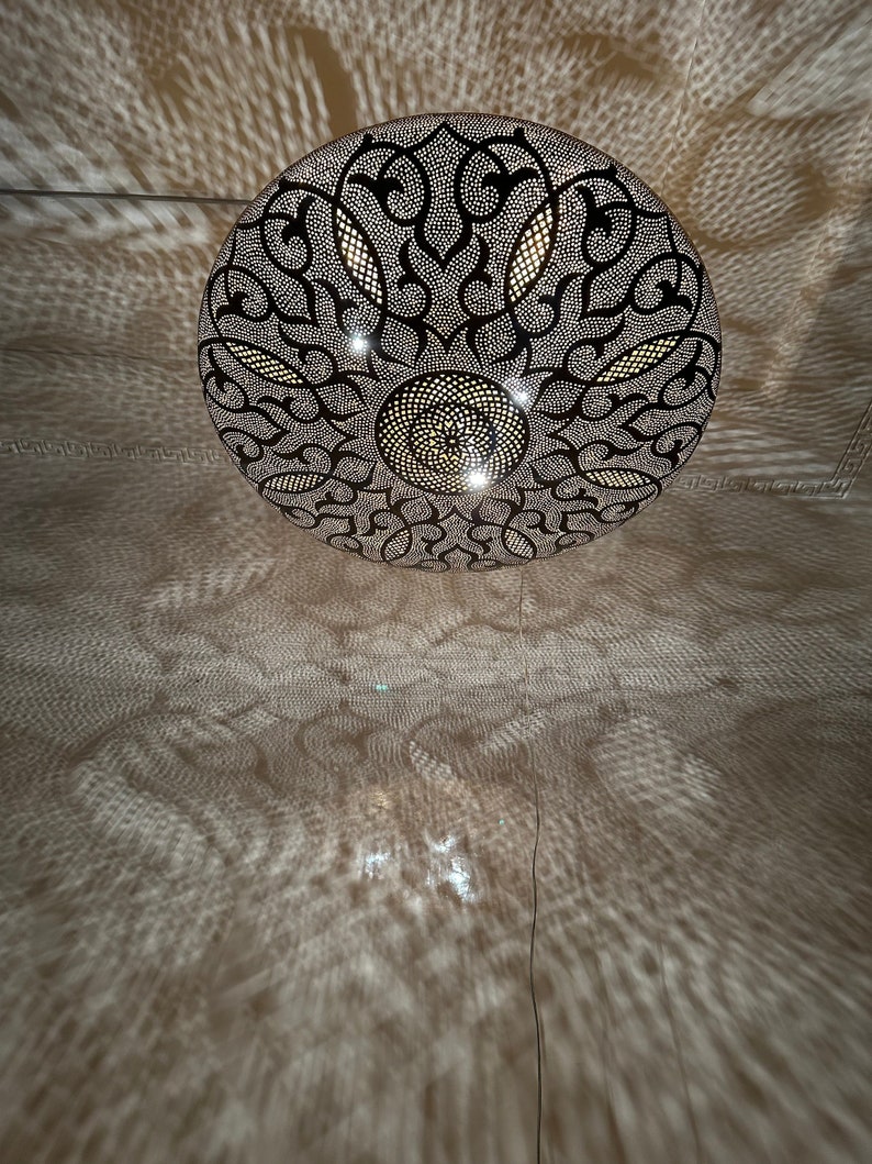 May include: A round, black metal ceiling light fixture with a decorative, intricate pattern. The light is on and casting a shadow on the ceiling.