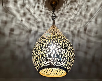 May include: A decorative pendant light with a teardrop shape. The metal shade features intricate cut-out patterns, casting ornate shadows on the ceiling. The light fixture is suspended by a chain and has a warm, inviting glow.