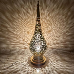 May include: A tall, gold-colored table lamp with a decorative, intricate, cut-out design. The lamp has a teardrop-shaped base and a pointed top.