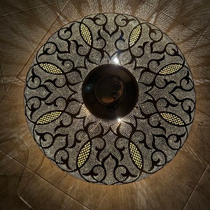 May include: A round, black metal ceiling light fixture with a decorative, intricate pattern. The light fixture is illuminated, casting a warm glow.