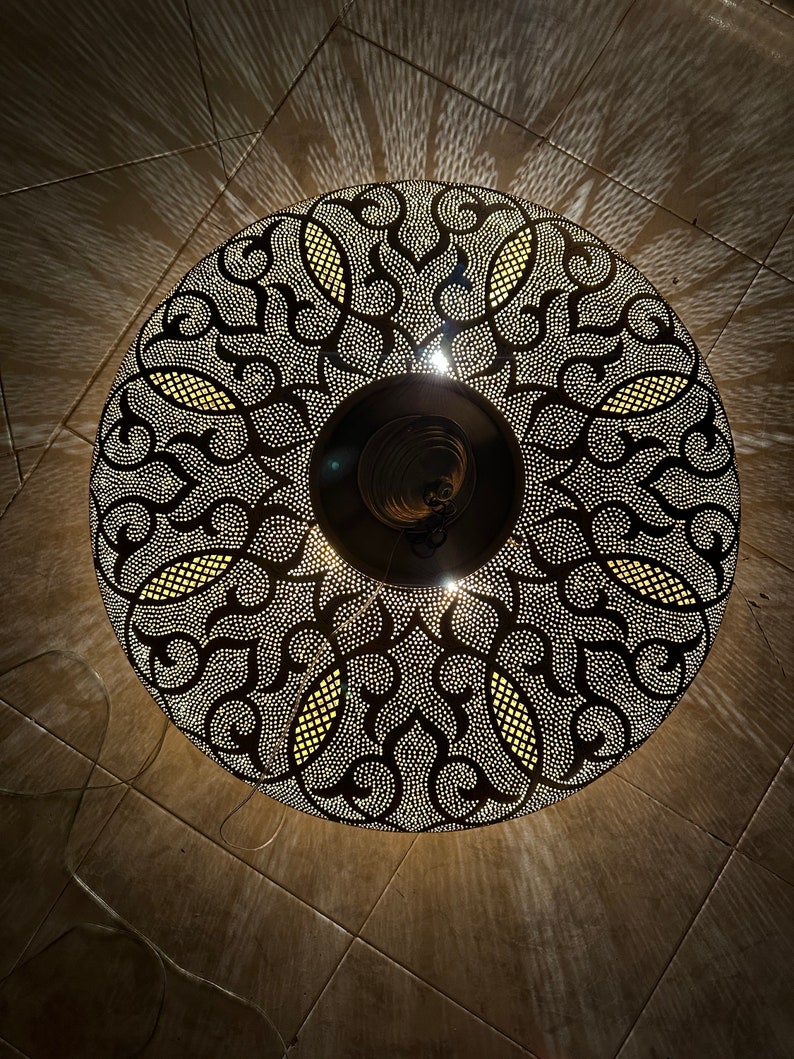 May include: A round, black metal ceiling light fixture with a decorative, intricate pattern. The light fixture is illuminated, casting a warm glow.