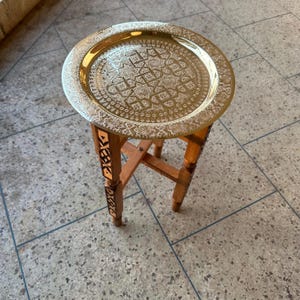 May include: A round gold-coloured metal tray table with an intricate geometric pattern engraved on the surface. The table features four wooden legs with inlaid decorative details, connected by a cross-brace structure for stability and support.