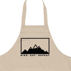 May include: A beige cotton apron with a large black pocket and adjustable straps. The apron has a black graphic of mountains with the text "HIKE - EAT - REPEAT" printed on the front.