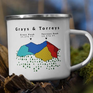 May include: White enamel camping mug with a colorful illustration of Grays & Torreys Peaks in Colorado. The mug has a silver rim and handle. The text "Grays Peak 14,270 FT." and "Torreys Peak 14,276 FT." is printed on the mug.
