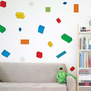 Op de afbeelding: Een witte muur met kleurrijke plastic blokken stickers in verschillende maten en kleuren. De stickers zijn in een willekeurig patroon gerangschikt. Op de voorgrond is een bank met een groen kikker knuffeldier erop.