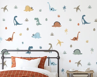 May include: A white wall with a variety of colorful dinosaur wall decals. The decals are in different sizes and shapes, and some have a cartoonish style. There is a bed with a brown and orange checkered blanket in the foreground.