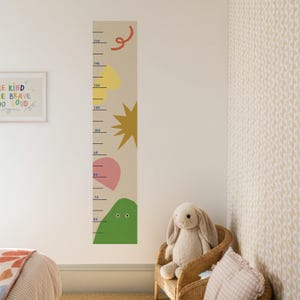 Puede incluir: Un medidor de altura beige con formas coloridas y medidas en centímetros. El medidor presenta un remolino rojo, un corazón amarillo, una estrella dorada, un corazón rosa y una forma de montaña verde. El medidor está montado en una pared blanca.