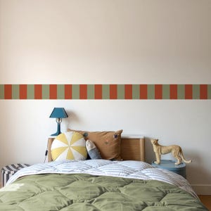 Könnte beinhalten: Ein Schlafzimmer mit einem Bett mit grün-weiß gestreifter Bettdecke, Kissen und einer dekorativen Bordüre aus abwechselnd roten und grünen vertikalen Streifen. Eine blaue Lampe und ein Spielzeugleopard sind ebenfalls zu sehen.