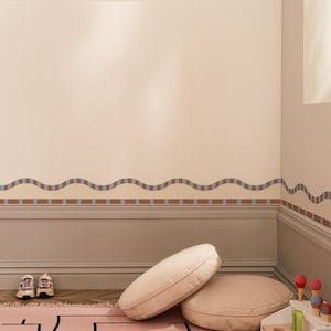 May include: A room with a beige wall featuring a wavy brown and blue border. Two round, light-coloured pillows rest on a pink rug. A pair of small trainers sits near the wall. The room has a neutral colour palette.