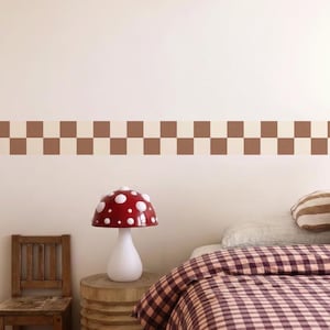 Op de afbeelding: Een slaapkamer met een rode paddenstoelvormige lamp met witte stippen, een houten stoel en een bed met een geruite dekbedovertrek. Een bruin en beige geruite rand siert de muur.