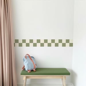 May include: A light blue backpack with orange and red accents sits on a green bench. The bench is made of wood and has a green cushion. Above the bench is a checkerboard pattern in green and off-white.