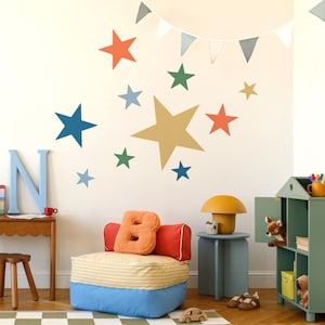 Peut inclure: Une chambre d'enfant avec des étoiles décoratives multicolores sur le mur, une lettre "N" bleue et un pouf coloré. Une petite table en bois et une armoire verte avec un cerf en jouet sont également visibles.
