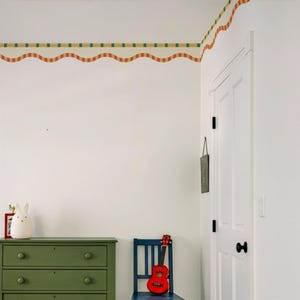 May include: A room corner with a white door, green dresser, and blue chair. A red ukulele rests on the chair. A decorative wall border features a wavy red and yellow pattern with green squares. A white bunny lamp sits on the dresser.