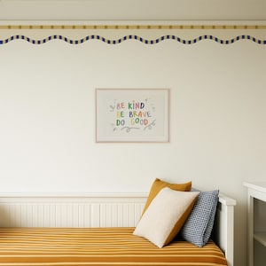 May include: A framed print with the words "BE KIND, BE BRAVE, DO GOOD" in colourful lettering hangs on a white wall. Below, a bed with a mustard-coloured striped cover is adorned with pillows in mustard, cream, and navy blue gingham.