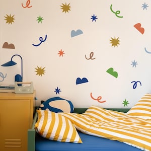 Puede incluir: Dormitorio infantil con cama azul, ropa de cama a rayas amarillas y mesita de noche amarilla. La pared blanca está decorada con pegatinas de estrellas, nubes y espirales de colores. Una lámpara azul y una pequeña radio están en la mesita.