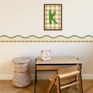 Op de afbeelding: Een kinderkamer met een houten bureau, stoel en een veelkleurige kruk. Een ingelijste print met de groene letter "K" hangt boven het bureau. Een digitale wekker geeft de tijd 10:15 aan.