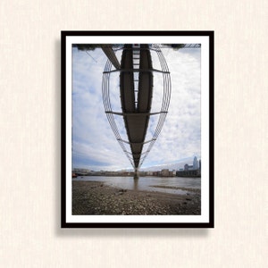 May include: Framed photograph of the Millennium Bridge in London, viewed from below. The bridge is grey with a modern design, suspended over the River Thames. The sky is cloudy, and buildings are visible in the background.