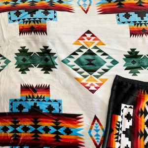 May include: A white fleece blanket with a Southwestern-inspired geometric pattern in shades of red, orange, yellow, blue, green, and black. The blanket has a black fleece backing.