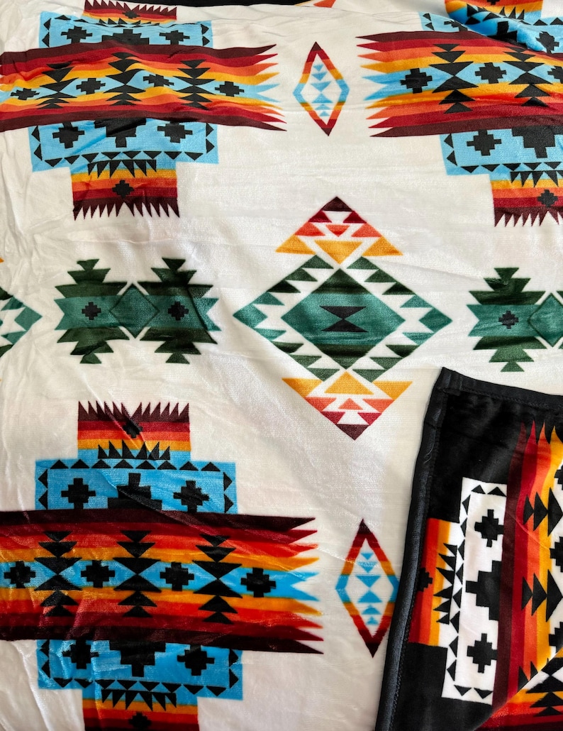 May include: A white fleece blanket with a Southwestern-inspired geometric pattern in shades of red, orange, yellow, blue, green, and black. The blanket has a black fleece backing.