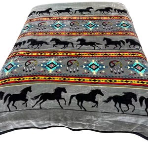 May include: A gray plush blanket featuring a repeating pattern of black running horses, dreamcatchers, and geometric shapes in red, yellow, and turquoise. The design is arranged in horizontal bands across the soft, textured fabric surface.