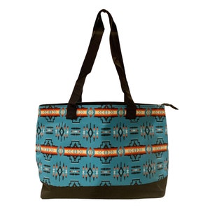 Pode incluir: Uma bolsa tote turquesa com um padrão geométrico em tons de laranja, vermelho, branco e preto. A bolsa tem alças pretas e fecho de correr.