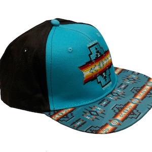 Op de afbeelding: Een turquoise en zwarte baseballpet met een Native American geïnspireerd ontwerp aan de voorkant en een gepatenteerde rand.