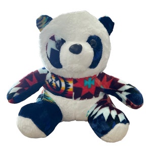May include: A white plush panda bear with black eyes and a black nose. The bear is wearing a colorful patterned fabric that includes red, blue, yellow, and white.